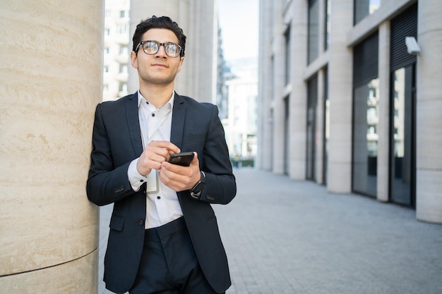 A man in a suit stands in front of a building and looks at his phone.