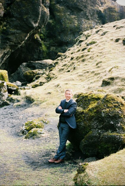 Man in a suit stands near a huge boulder covered with moss Iceland