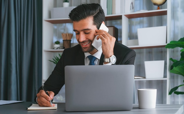 A man in a suit talks on a phone while sitting at a desk with a laptop.