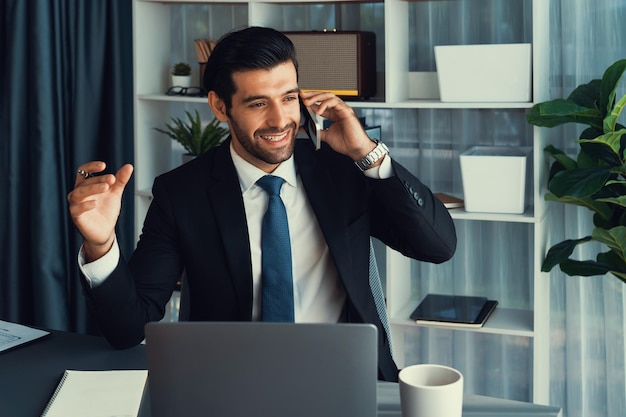 A man in a suit talks on a phone while sitting at a desk.