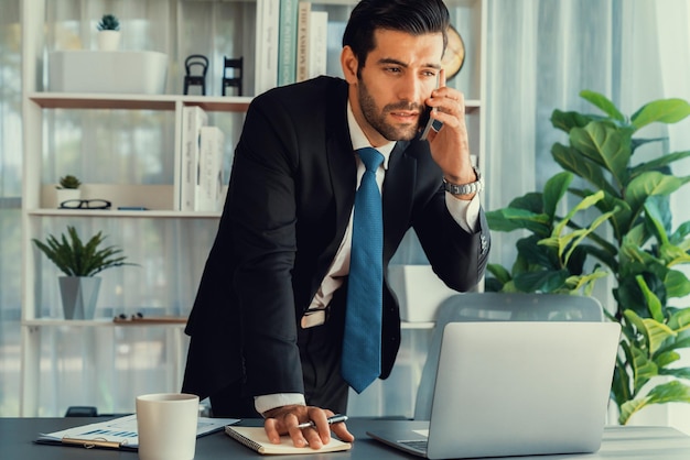 A man in a suit talks on a phone while standing in front of a laptop.