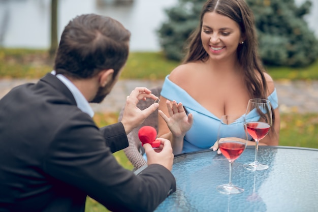 Man in suit with his back to camera putting ring on finger of his beloved happy woman sitting at table outdoors