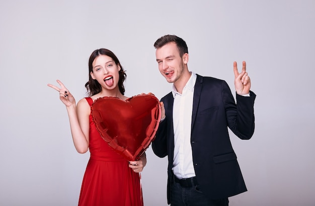 Man in suit and woman in red dress hold a red heart shaped ballon do peace sign on white background