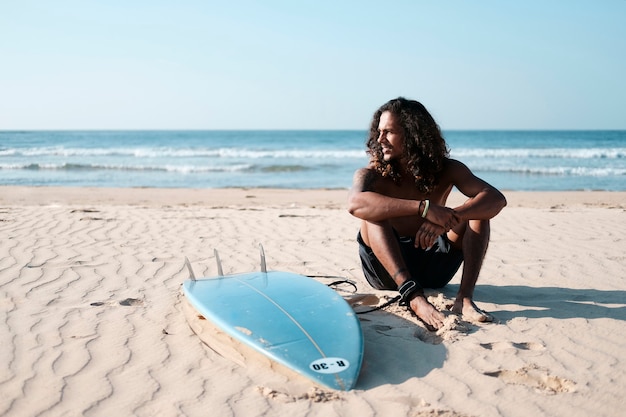 Man Surfer Sitting at Surfboard on Sand Beach