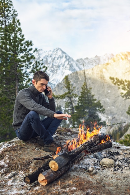 Man in a sweater talking on the phone near a campfire