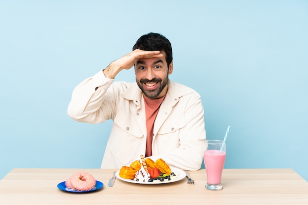 Man at a table having breakfast waffles and a milkshake looking far away with hand to look something