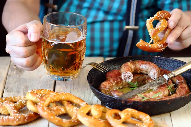 A man at the table with a glass of beer, a pretzel and a frying pan with sausages. Oktoberfest celebration.