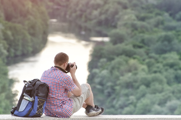男は森と川の背景にある丘から写真を撮ります