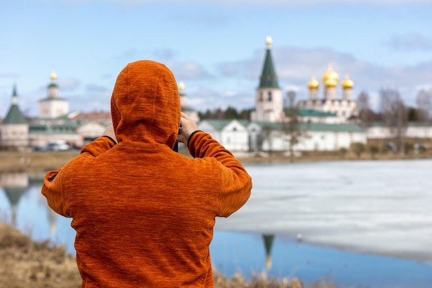 Мужчина фотографирует на свой телефон старый древний монастырь, достопримечательность и озеро, тающее от снега в весенний день.