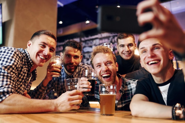 Man takes selfie by phone group of people together indoors in\
the pub have fun at weekend time