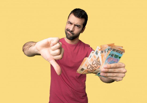 Man taking a lot of money showing thumb down with both hands on isolated yellow background