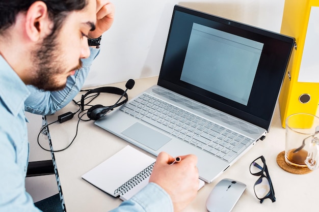 Man taking notes in notebook while using laptop at home Young freelancer writing details on book