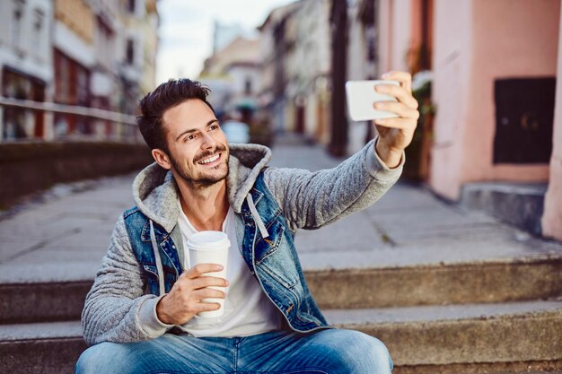 Man taking selfie holding coffee and sitting on stairs