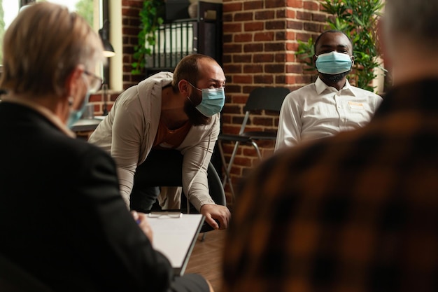 Man talking to people at aa group meeting about mental health,\
having face mask. multi ethnic patients with addiction attending\
therapy session and having conversation about problems.