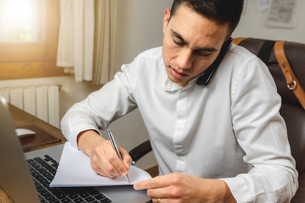 man talking to the phone while taking note and working on computer.