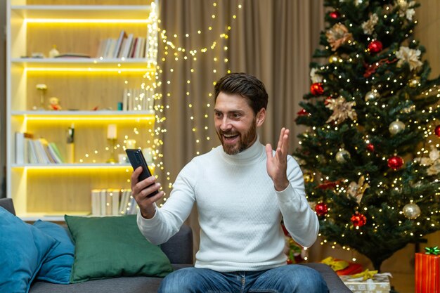 A man talks on a video call at home with a phone sits on a sofa\
near a christmas tree records video