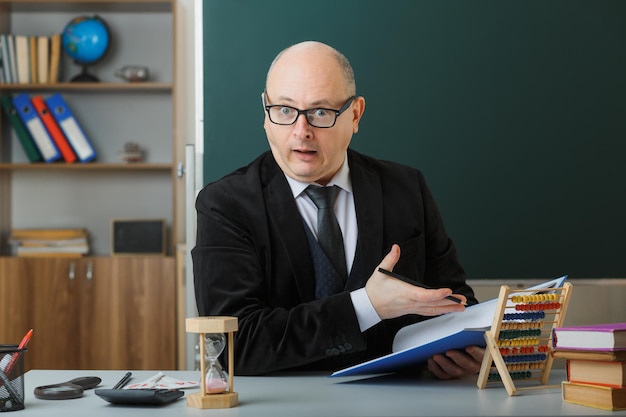 Man teacher wearing glasses checking class register looking confused and surprised sitting at school desk in front of blackboard in classroom