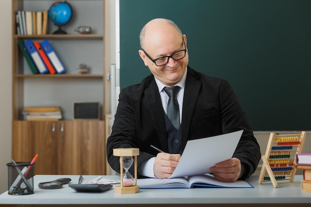 Man teacher wearing glasses sitting at school desk in front of blackboard in classroom checking homework of students happy and pleased smiling