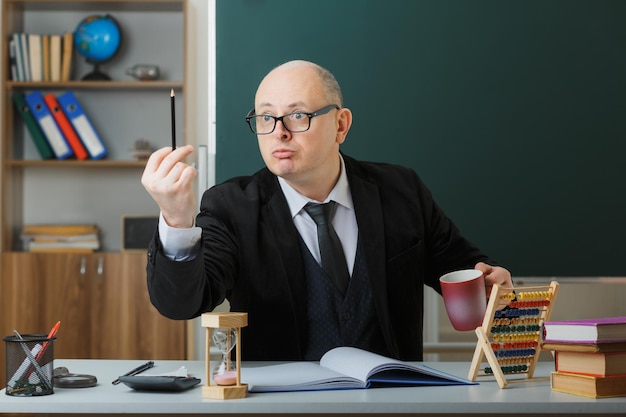 man teacher wearing glasses sitting at school desk in front of blackboard in classroom holding mug of coffee explaining lesson