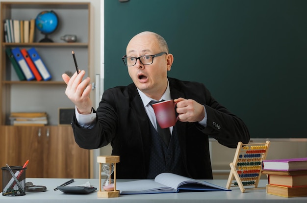 Man teacher wearing glasses sitting at school desk in front of blackboard in classroom holding mug of coffee looking aside surprised