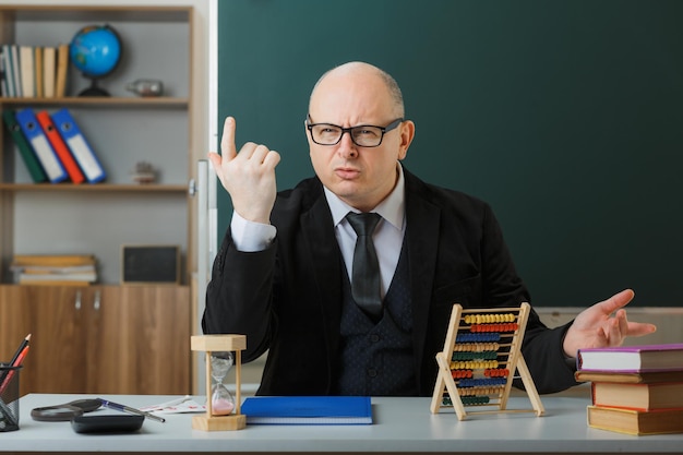 Man teacher wearing glasses sitting at school desk with class register in front of blackboard in classroom explaining lesson being displeased gesturing with hands