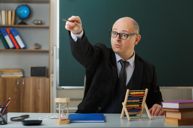 Man teacher wearing glasses sitting at school desk with class register in front of blackboard in classroom explaining lesson looking surprised