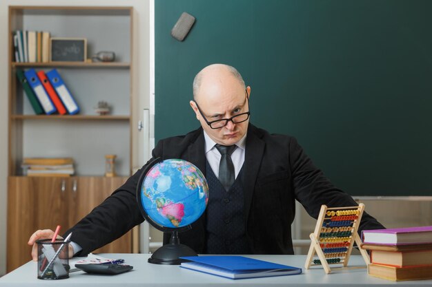 Man teacher wearing glasses sitting with globe at school desk in front of blackboard in classroom explaining lesson looking frowningly