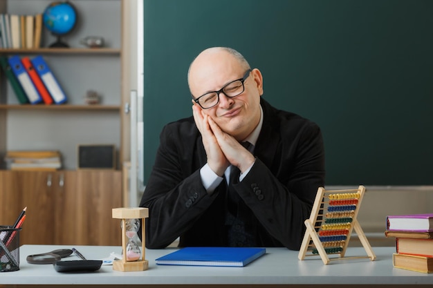 Man teacher wearing glasses with class register sitting at school desk in front of blackboard in classroom looking tired and bored wants to sleep leaning head on palms