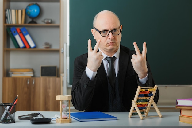 Man teacher wearing glasses with class register sitting at school desk in front of blackboard in classroom using abacus showing number four with fingers