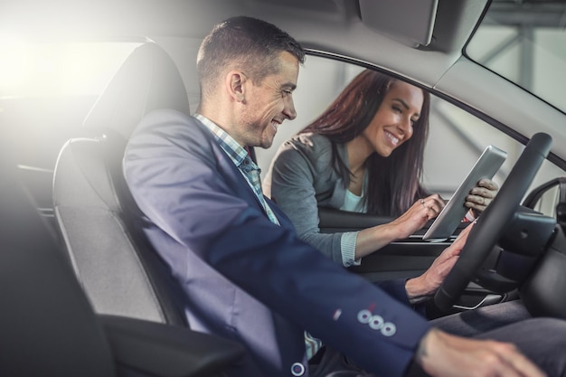 販売店で車をテストしている男性が、技術を示している女性の自動車ディーラーの手でタブレットを見て微笑んでいます
