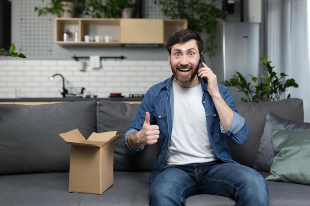 Man thuis ontving een bestelling van de online winkel blij opent een kartonnen doos met een pakket laat een positieve reactie achter via de telefoon