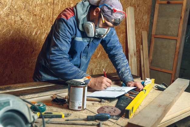 Man timmerman in zijn thuisstudio werken met hout en tekent schetsen in potlood in een notitieblok