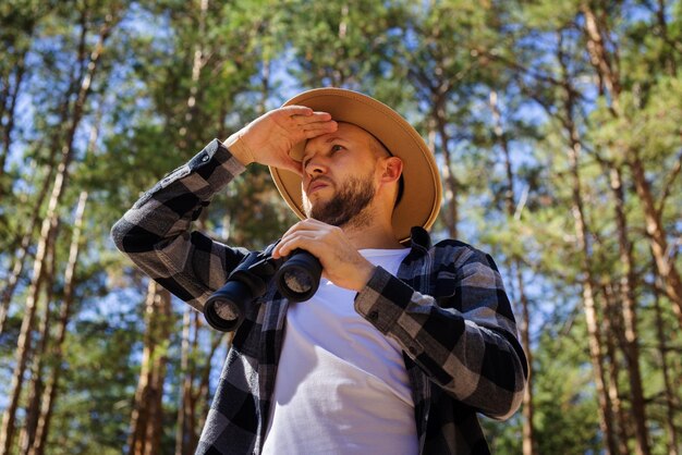 Man tourist in a hat and a plaid shirt looks through binoculars in the forest