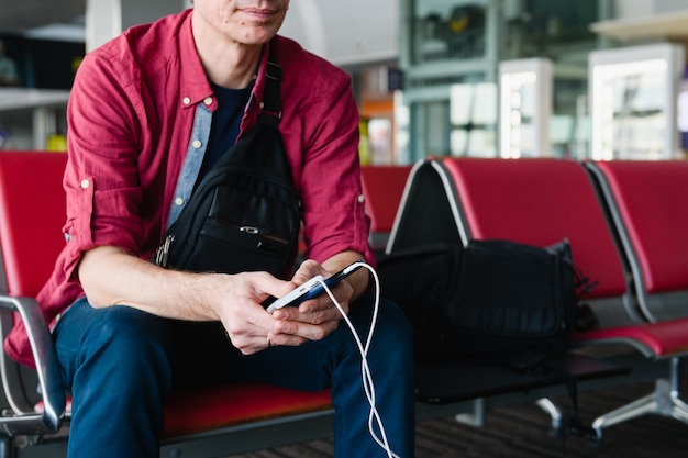 Foto l'uomo turistico tiene lo smartphone in carica tramite cavo usb dalla banca di alimentazione portatile, seduto con i bagagli al cancello dell'aeroporto