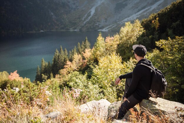 남자 관광객은 언덕 꼭대기에서 휴식을 취하며 산과 호수의 멋진 풍경을 바라보고 있습니다.