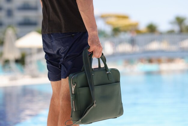 ショートパンツの男の観光客はホテルのプールの背景にビジネスバッグを保持します