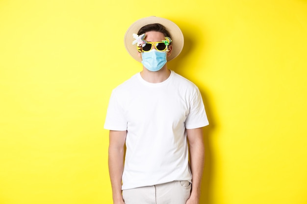 Man tourist wearing medical mask and summer hat with sunglasses, going on trip during pandemic, yellow wall