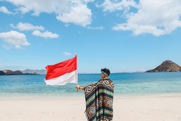 Un uomo in tessuto tradizionale che sventola la bandiera dell'indonesia sulla spiaggia