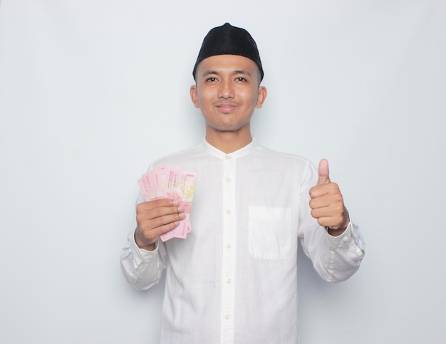 A man in a traditional malay outfit holds a bunch of pink money and shows thumbs up.