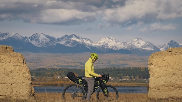 남자는 자전거 포장과 함께 혼합 지형 사이클 여행을 여행합니다. 자전거 가방을 들고 여행하는 여행자 녹색 검정 색상의 스포츠 자전거 포장 자전거 운동복 산 눈 덮인 돌 아치