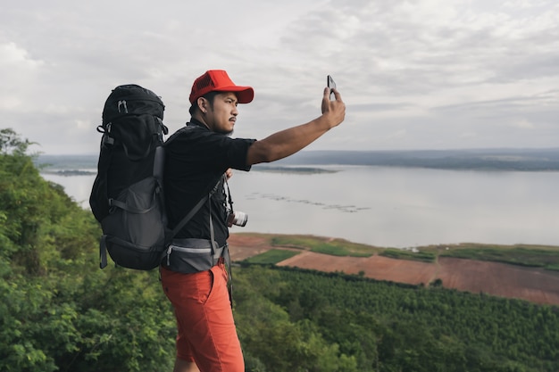 Man traveler with backpack using smartphone take a selfie on edge of cliff, on a top of the rock mountain