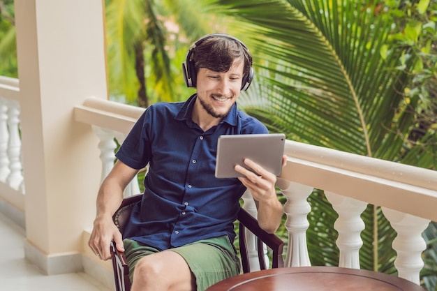 Man in the tropics talking with friends and family on video call using a tablet and wireless headphones sitting on the terrace.