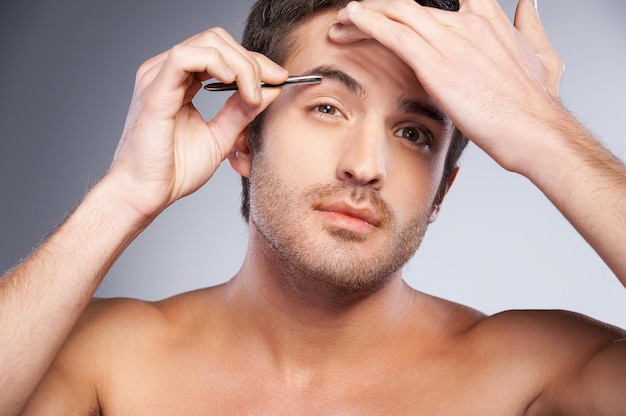 Man tweezing his eyebrows. Handsome young man tweezing his eyebrows and looking at camera while standing isolated on grey background