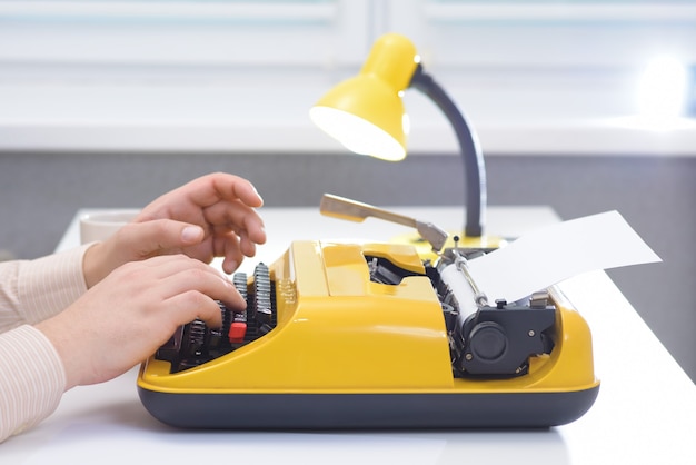 Man typing on yellow typewriter with lamp on white office desk near the window