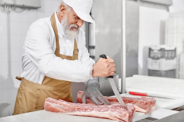 Man in uniform, apron cutting meat with knife on factory.