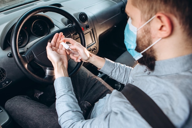 男は車を運転中に消毒剤を使用します。コロナウイルス流行時の注意事項。車で医療マスクの男。