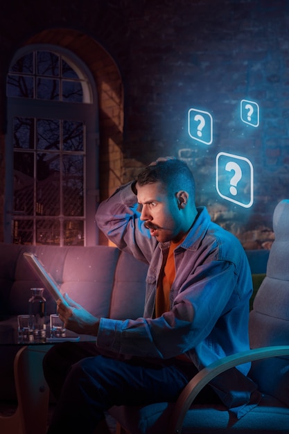 Uomo che usa gadget e riceve notifiche al neon a casa di notte. seduti in poltrona, navigando in internet e cercando informazioni. abuso dei social media, chat e navigazione, dipendenza da gadget.