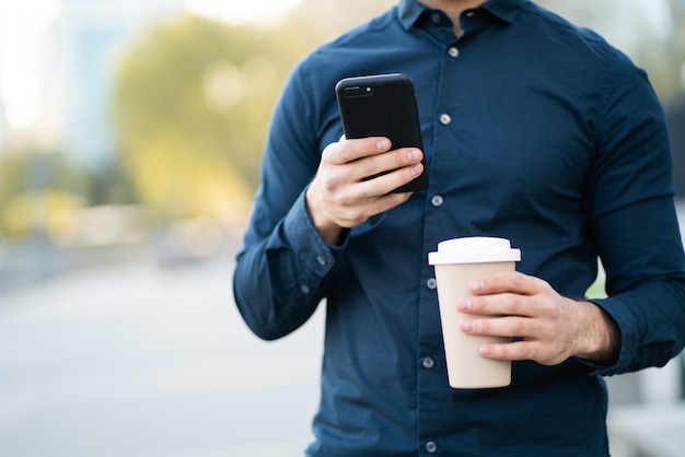 Man using his mobile phone and holding a cup of coffee while standing outdoors at the street