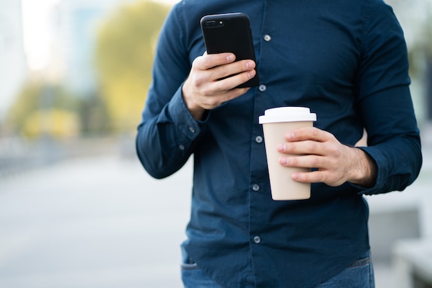 Uomo che utilizza il suo telefono cellulare e che tiene una tazza di caffè mentre si trovava all'aperto in strada