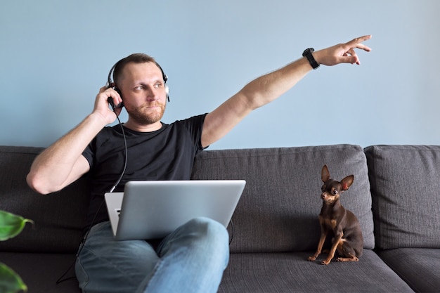 Foto uomo che usa il laptop a casa, maschio che si rilassa seduto sul divano con le cuffie che ascolta e si gode musica, riposo e tempo libero, sfondo domestico